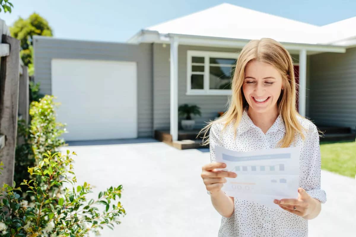 a woman stands outside a modern house, holding a document with a mix of pride and anticipation