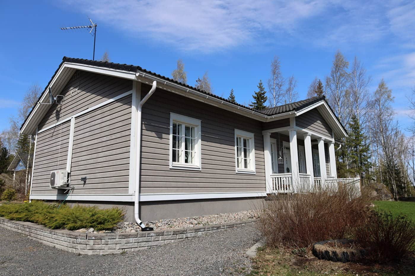Moderni talon julkisivu, jossa on pieni puinen kuisti, lämpöpumppuyksikkö ja sorapolku, kirkkaan sinisen taivaan alla.