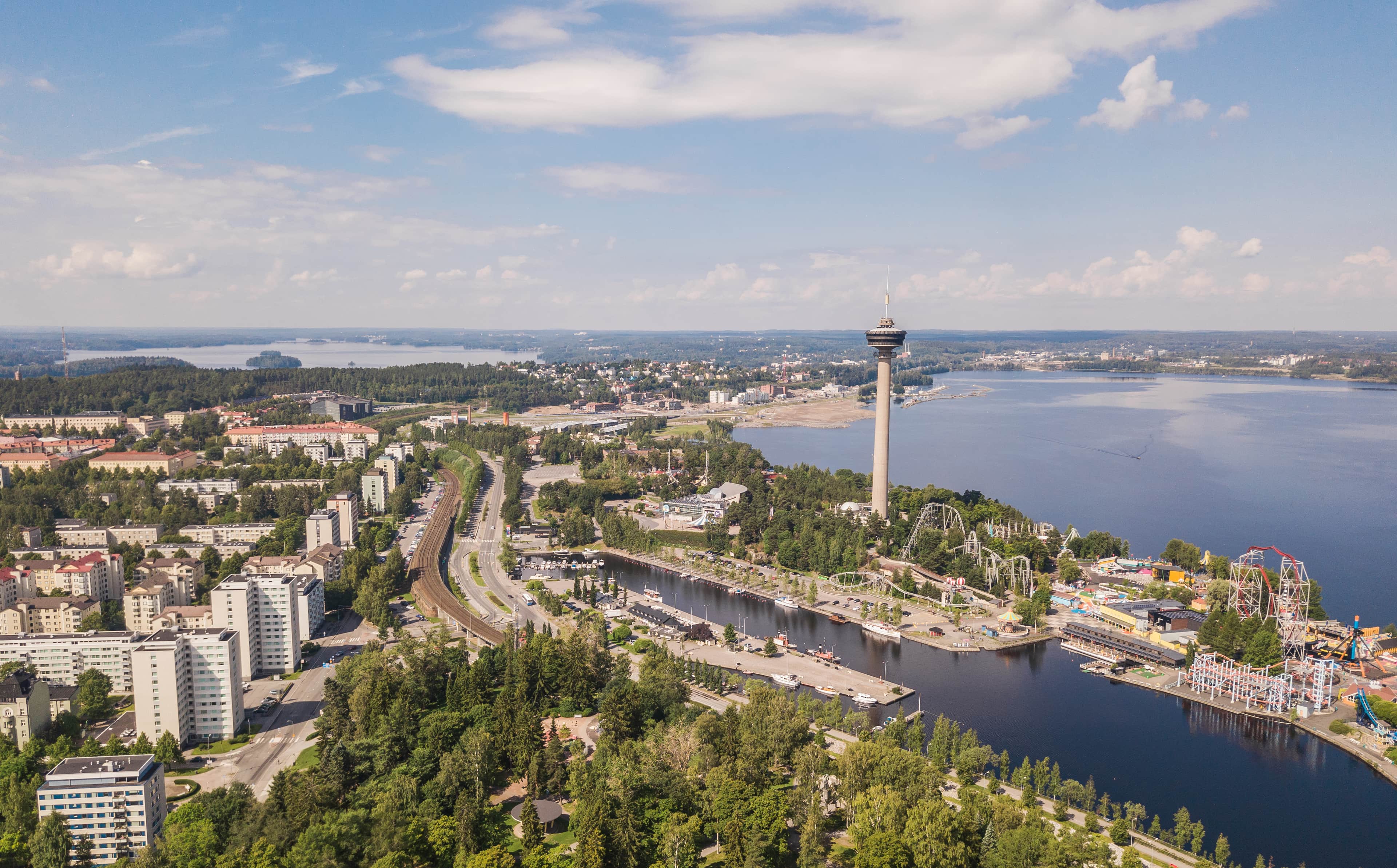 Ilmakuva Tampereen kaupungista, jossa näkyy veden äärellä sijaitseva näkötorni, huvipuisto, venesatama, mutkittelevia teitä ja asuinrakennuksia vihreiden puiden keskellä.