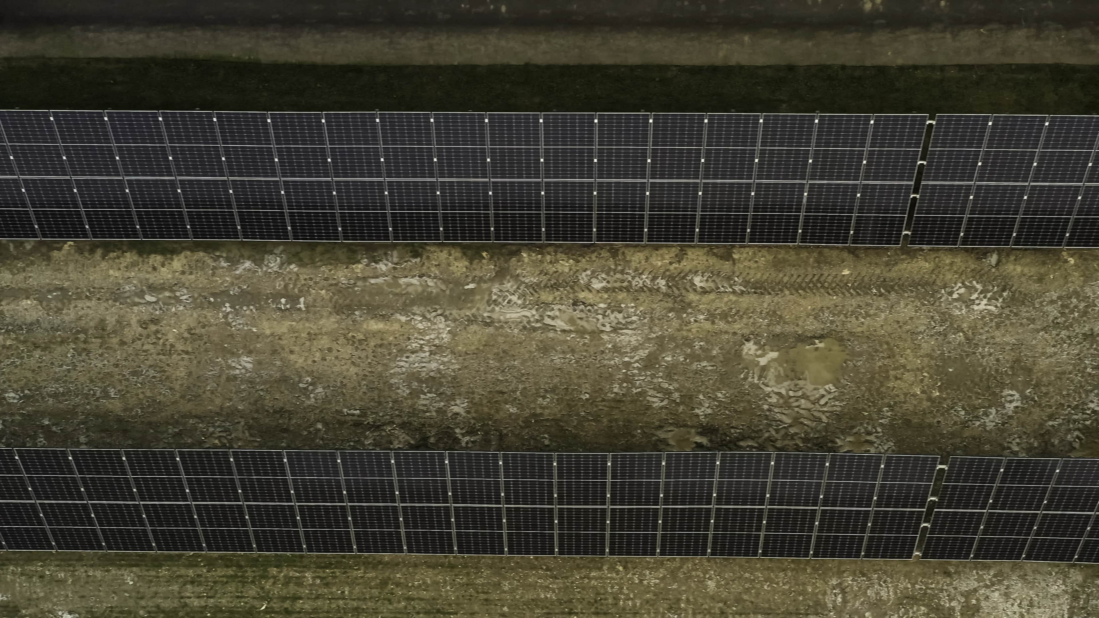 Aerial top-down view of ground-mounted solar panels arranged in parallel rows, with dirt and grass paths visible between the panel arrays.