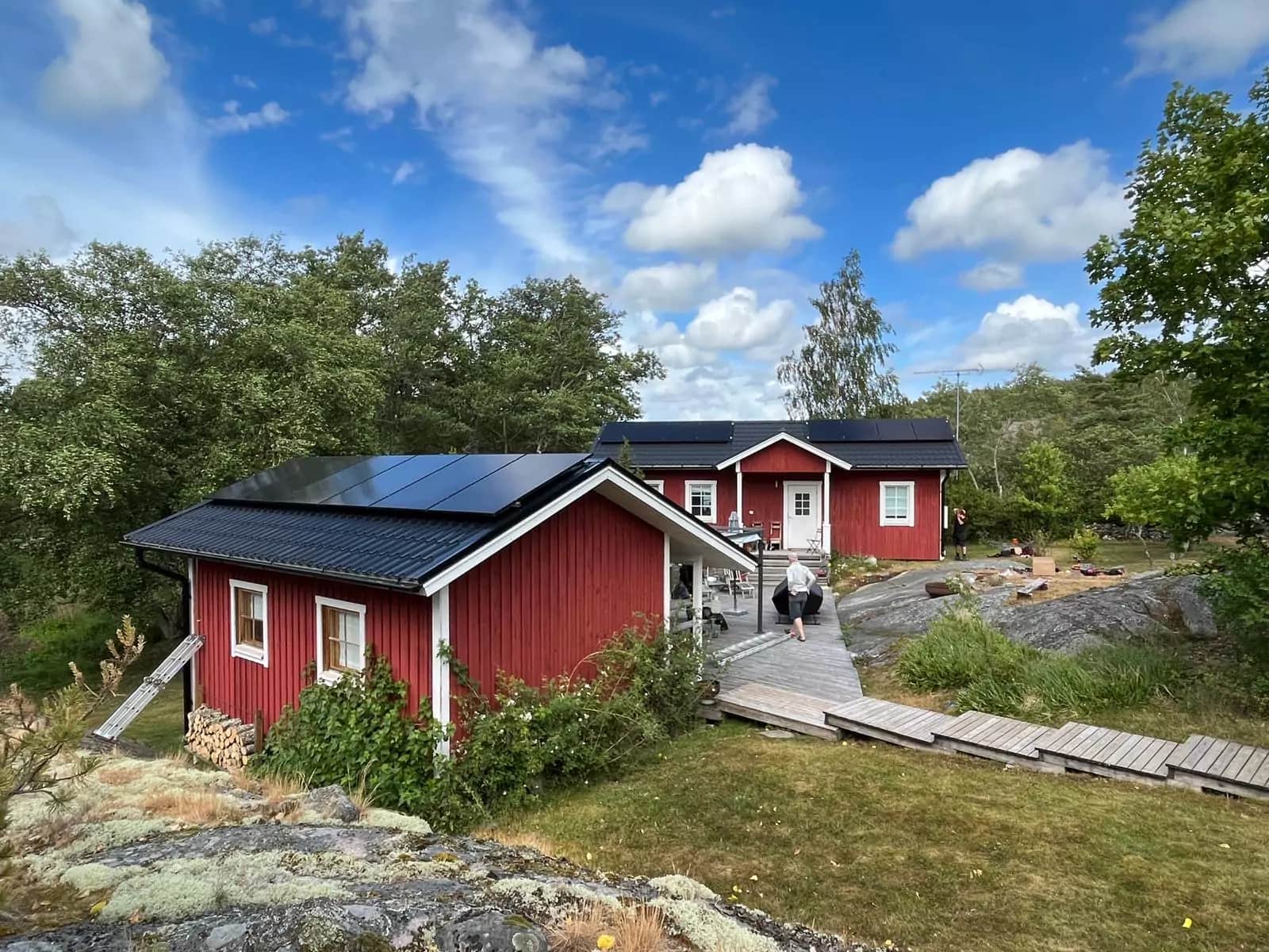 Punainen puutalo aurinkopaneeleilla, puiden ja kivisen maaston ympäröimänä osittain pilvisen taivaan alla. Henkilö kävelee puisella polulla.